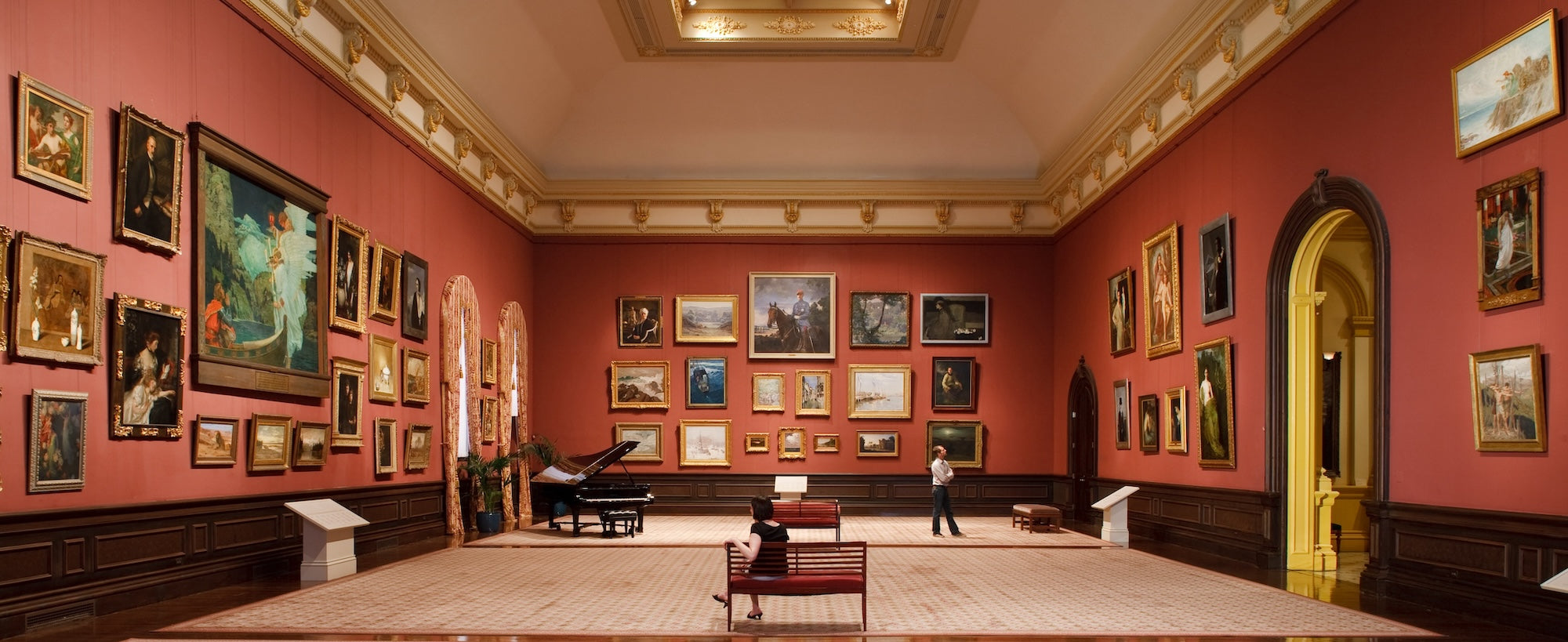 Very large art gallery room with salon style displays of about 50 paintings across three walls, all hung on picture rail molding hangers with adjustable hooks
