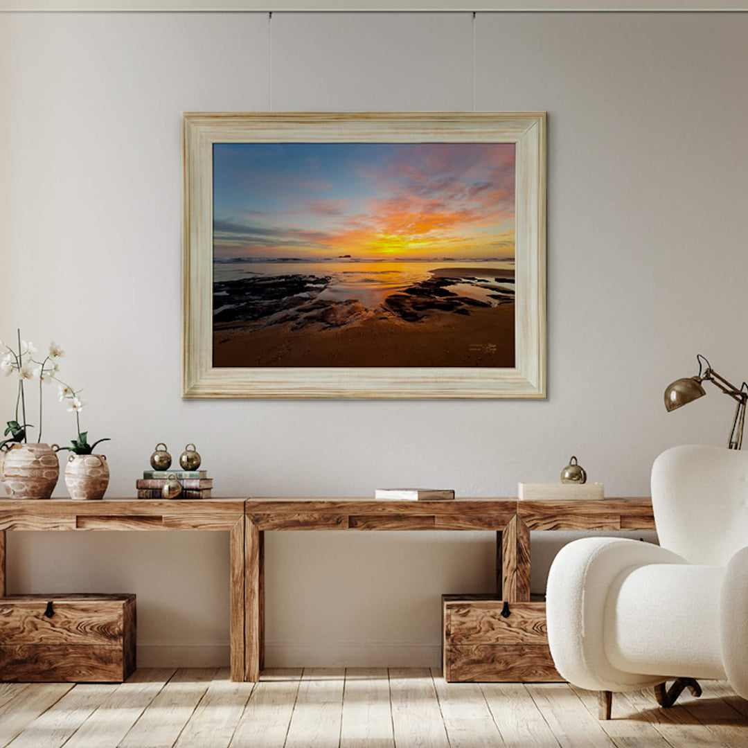 Framed artwork of a sunset over water in a living room setting with a wooden console table and white armchair.