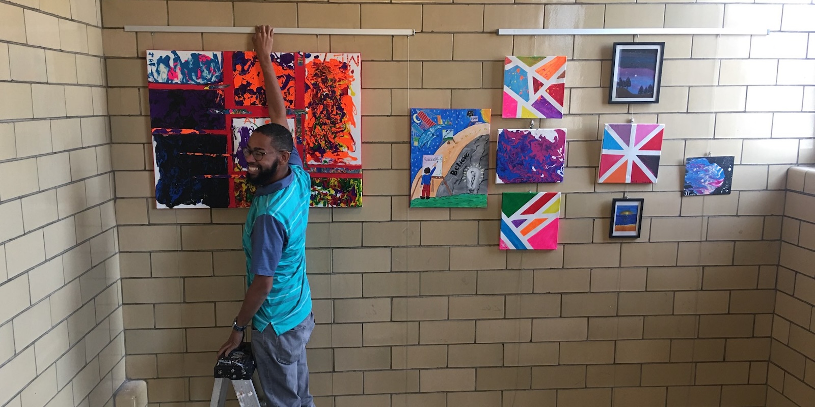 Teacher on ladder hanging student art in stairwell, using art hanging system